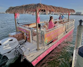 Pink tiki-themed pontoon boat with a thatched roof and leis, docked at a sunny coastal marina with passengers in pink outfits enjoying drinks on the water