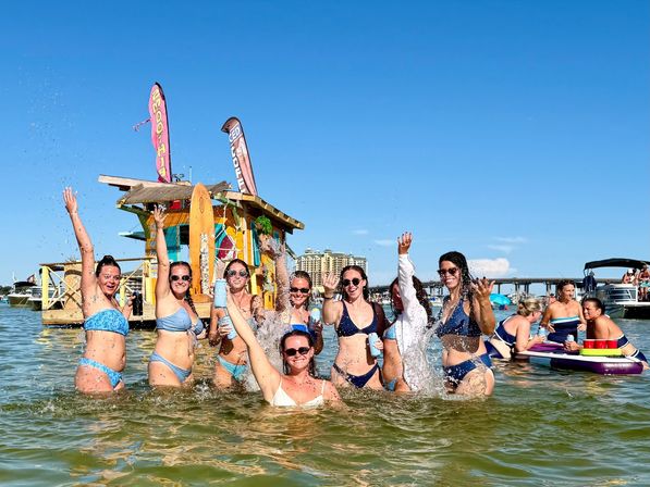 Group of friends in bikinis splashing and cheering in shallow coastal water beside a colorful floating tiki shack and nearby boats on a sunny blue-sky summer day