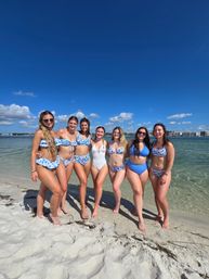 Seven friends in blue-and-white swimsuits smiling and posing on a sunny sandy beach beside calm turquoise water under a bright blue sky with fluffy clouds.