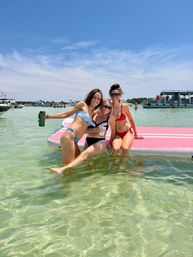 Three friends in bikinis laughing and posing on a pink inflatable dock in shallow clear turquoise water, holding drinks with boats and a sunny blue sky in the background — lively summer beach scene.