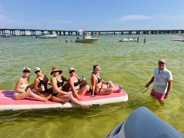 Group of friends lounging on a pink inflatable floating mat in shallow green coastal bay water, sipping drinks with anchored boats, jet skis and a long bridge on a sunny summer beach day