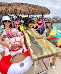 Group of friends on a tiki-top pontoon boat at a marina, wearing colorful retro outfits and bucket hats, gathered around a resin bar with an inflatable doll and life-ring float.