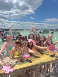 Energetic group of women in colorful swimsuits cheering at a crowded beach boat party in shallow turquoise water with pontoon boats and slides under a bright blue sky with fluffy clouds.