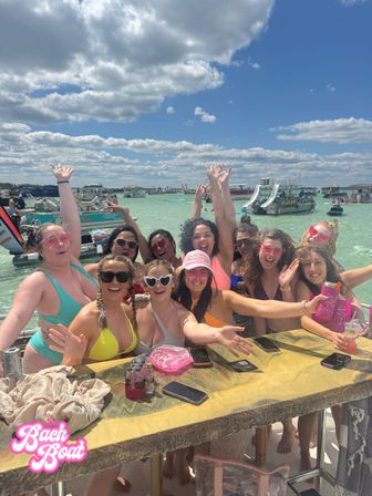 Energetic group of women in colorful swimsuits cheering at a crowded beach boat party in shallow turquoise water with pontoon boats and slides under a bright blue sky with fluffy clouds.