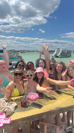 Energetic group of women in colorful swimsuits cheering at a crowded beach boat party in shallow turquoise water with pontoon boats and slides under a bright blue sky with fluffy clouds.