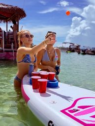 Two people in bikinis playing beer-pong on a paddleboard in clear shallow tropical water with red cups and drinks on the board, tiki hut and boats nearby under a sunny blue sky — lively beach day vibe