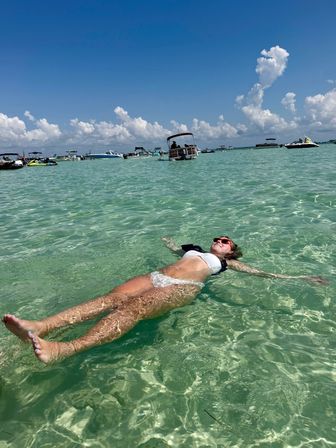 Person in a white bikini and red sunglasses floating on their back in crystal-clear turquoise water, anchored boats on the horizon under a bright blue sky with puffy clouds