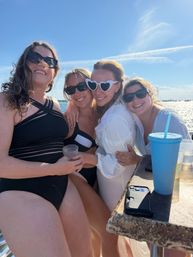 Four smiling women in swimsuits and sunglasses hugging on a sunny boat cruise over sparkling coastal water, blue sky, with a blue tumbler and phone on the railing.