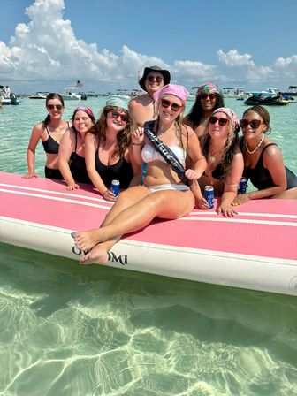 Smiling group of friends in swimsuits lounging on a large pink inflatable mat in clear turquoise shallow water with boats and jet skis anchored nearby under a sunny blue sky, festive celebration vibe.