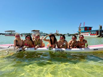 Eight friends lounging on a large pink-and-white paddleboard in shallow clear green water at a sunny coastal sandbar, wearing swimsuits and pink sunglasses, holding drinks with tiki boats and colorful floating food shacks visible in the background.