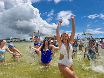 Beach party on a sunny sandbar — a group of women in swimsuits splashing and cheering in shallow green water with boats and a tiki hut under a blue sky and dramatic clouds.