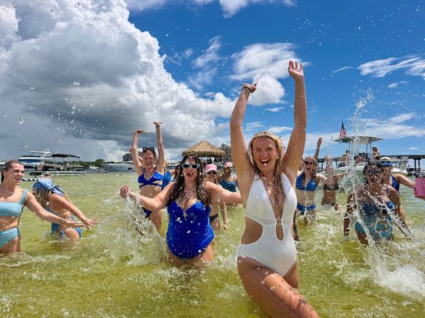 Beach party on a sunny sandbar — a group of women in swimsuits splashing and cheering in shallow green water with boats and a tiki hut under a blue sky and dramatic clouds.