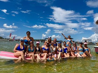 Group of friends in colorful bikinis enjoying a sunny sandbar party on a pink floating mat in shallow green water, surrounded by anchored boats under a bright blue sky with puffy clouds