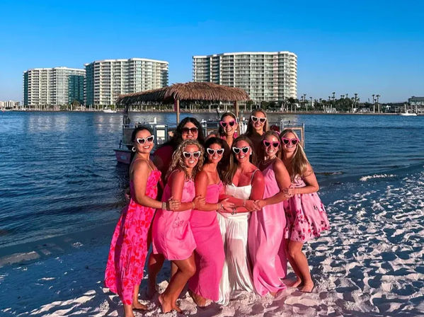 Smiling group of women arm-in-arm wearing pink dresses and heart-shaped sunglasses, one in white, on a sunny sandy beach by a marina with a tiki-style boat and waterfront high-rise condos