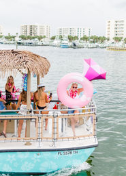 Women partying on a tiki‑roof pontoon boat on Florida waterfront, wearing leis and swimsuits, one holding a big pink diamond‑shaped inflatable, marina and condos in the background