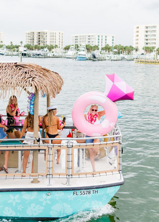 Women partying on a tiki‑roof pontoon boat on Florida waterfront, wearing leis and swimsuits, one holding a big pink diamond‑shaped inflatable, marina and condos in the background