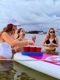 Laughing women in swimsuits playing floating beer-pong with red cups on a paddleboard in shallow bay water, holding canned drinks with boats and a cloudy sky in the background