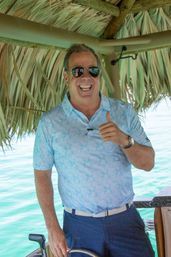 Smiling man in sunglasses giving a thumbs-up at a boat helm under a thatched tiki roof, wearing a light blue floral polo and navy shorts with turquoise tropical water in the background