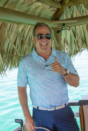 Smiling man in sunglasses giving a thumbs-up at a boat helm under a thatched tiki roof, wearing a light blue floral polo and navy shorts with turquoise tropical water in the background