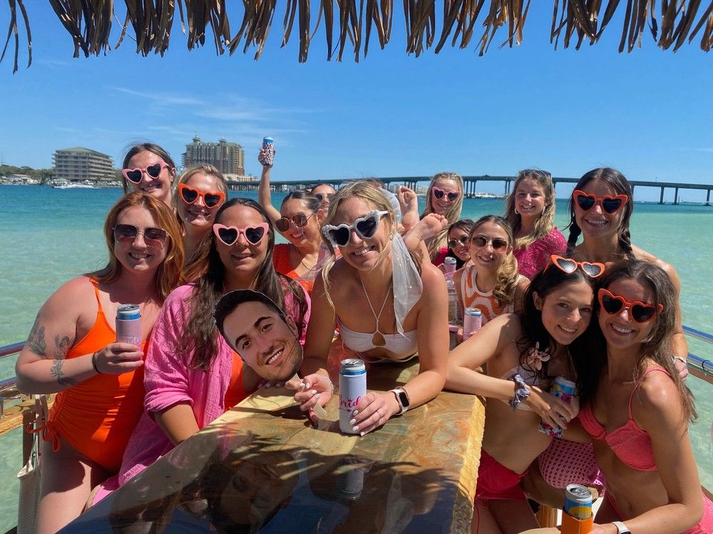 Colorful group of women in swimsuits and heart-shaped sunglasses enjoying a sunny boat party under a thatched tiki roof, holding canned drinks with turquoise water, a bridge, and beachfront buildings in the background.