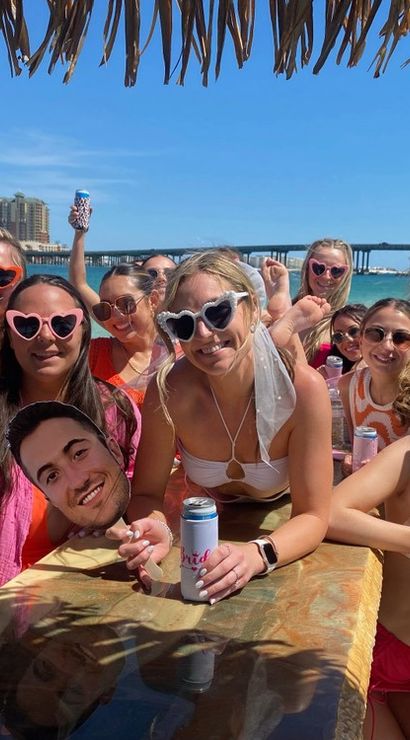 Colorful group of women in swimsuits and heart-shaped sunglasses enjoying a sunny boat party under a thatched tiki roof, holding canned drinks with turquoise water, a bridge, and beachfront buildings in the background.