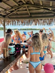 Group of friends in colorful bikinis enjoying drinks under a thatched tiki roof on a docked boat, with calm blue bay water and a tree-lined shoreline in the background.