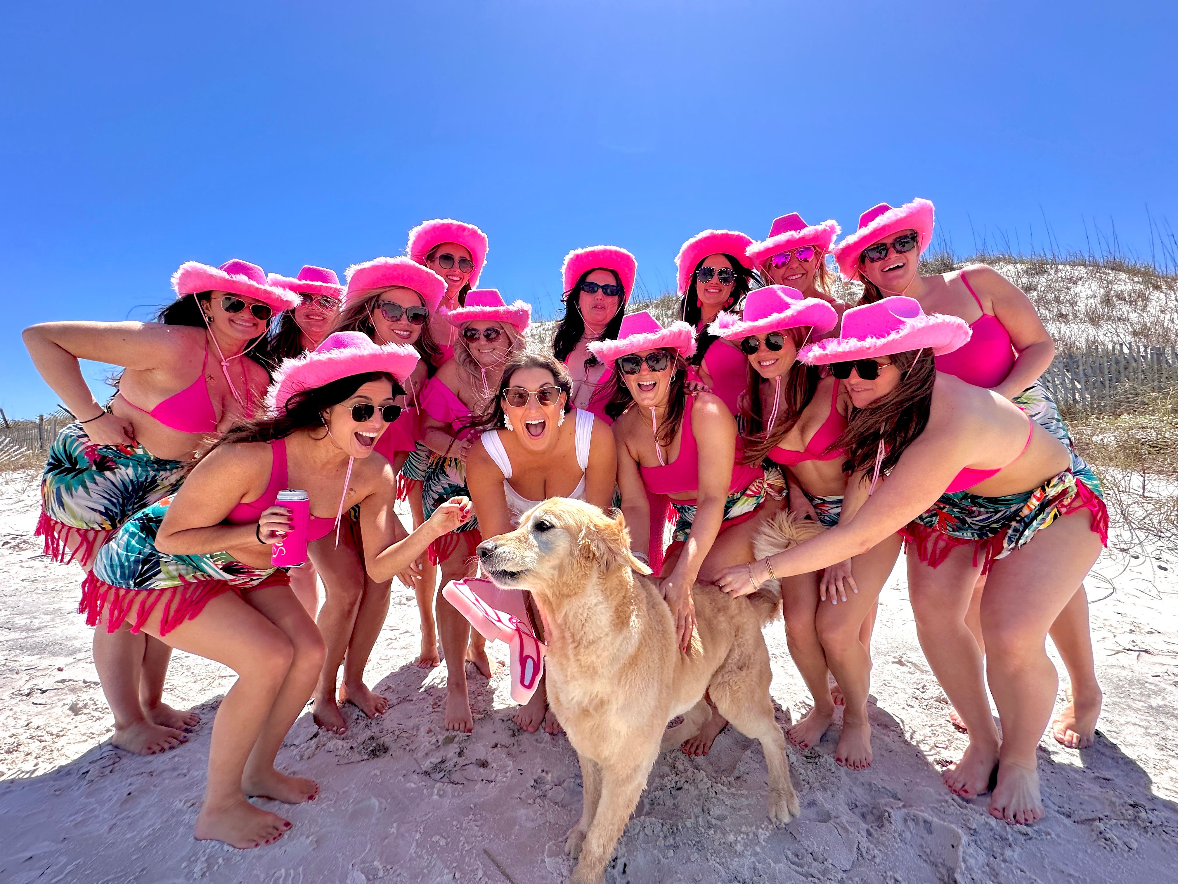 Sunny white-sand beach group of women in matching pink cowboy hats, pink bikinis and tropical sarongs, smiling in sunglasses around a golden retriever wearing a pink bandana and holding a flip-flop under a bright blue sky.