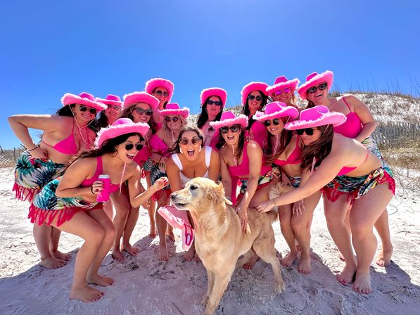 Sunny white-sand beach group of women in matching pink cowboy hats, pink bikinis and tropical sarongs, smiling in sunglasses around a golden retriever wearing a pink bandana and holding a flip-flop under a bright blue sky.