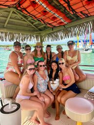 Ten friends in colorful swimsuits smiling under a thatched tiki roof on a boat party over clear turquoise water, with bright orange life jackets overhead and festive flags and boats in the background.