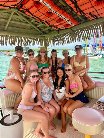 Ten friends in colorful swimsuits smiling under a thatched tiki roof on a boat party over clear turquoise water, with bright orange life jackets overhead and festive flags and boats in the background.