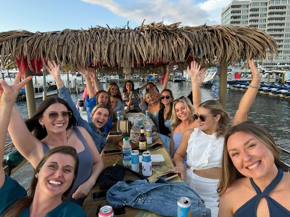 Smiling group of women on a tiki-style party boat at a marina, raising hands and enjoying drinks at a long table with waterfront condos in the background.