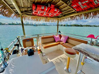Tiki-roof pontoon boat lounge on turquoise bay with tan leather seating, pink pillows, a bar stool and distant city skyline on a sunny day.