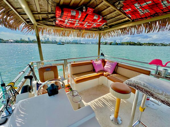 Tiki-roof pontoon boat lounge on turquoise bay with tan leather seating, pink pillows, a bar stool and distant city skyline on a sunny day.