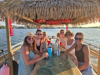 Group of friends smiling under a thatched tiki roof on a pontoon boat, drinks on the table as they cruise a sunlit waterfront inlet with a tropical, golden-hour vibe