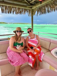 Two smiling women in sunglasses and colorful striped beach towels sitting under a thatched palapa roof on a boat, with bright turquoise tropical water and a distant shoreline in the background.