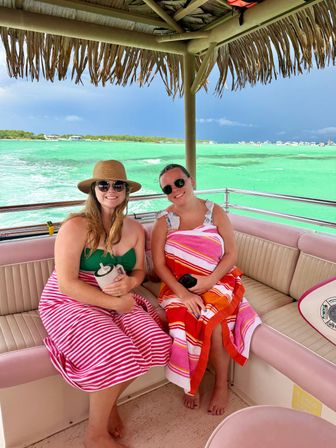 Two smiling women in sunglasses and colorful striped beach towels sitting under a thatched palapa roof on a boat, with bright turquoise tropical water and a distant shoreline in the background.