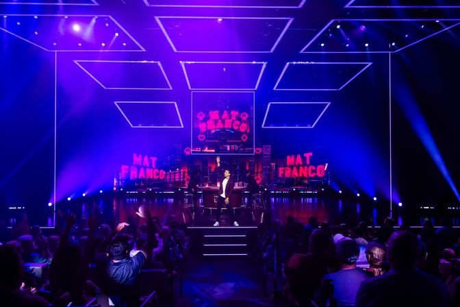 Solo performer on a brightly lit indoor theater stage under purple neon lights and geometric LED ceiling panels, cityscape backdrop and cheering audience.