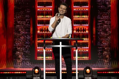 Smiling performer on a brightly lit theater stage doing a cups-and-balls magic trick at a tall round table with a colorful bottle-lined backdrop and spotlights