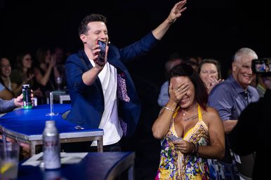 Live indoor entertainment scene: energetic host holding up a phone and raising his arm while a woman in a colorful dress covers her face laughing in surprised delight, audience clapping in the background.