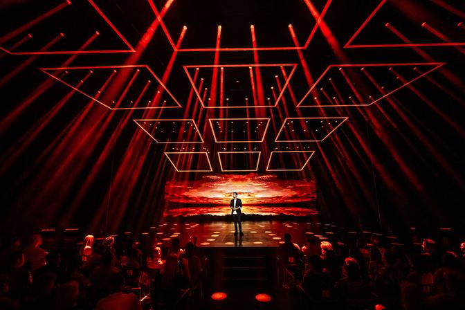 Solo performer on a dramatic indoor stage bathed in red spotlights, geometric illuminated ceiling frames and a fiery sunset LED backdrop, with silhouetted audience watching.