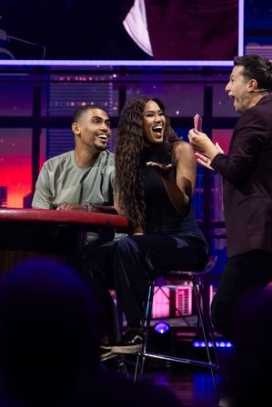 Lively studio stage scene with a magician showing playing cards to a seated couple; the woman laughs and the man looks amazed under purple city-skyline lighting, live show atmosphere.