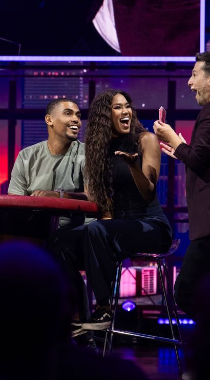 Lively studio stage scene with a magician showing playing cards to a seated couple; the woman laughs and the man looks amazed under purple city-skyline lighting, live show atmosphere.