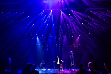 Stage performer with arms outstretched on a theater stage under dramatic blue and purple spotlights, mist and audience silhouettes during a live indoor show.