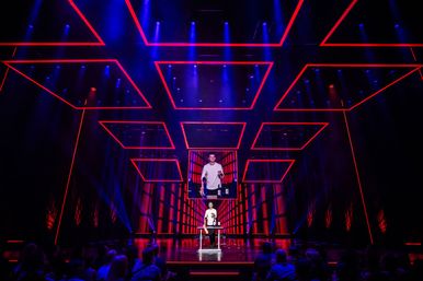 Neon-lit theater stage with a presenter at a table beneath red rectangular ceiling frames and blue spotlights, a large screen showing the speaker, and a seated audience watching the live event.