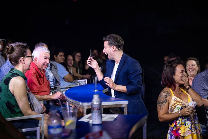 Live magic show at an indoor venue — a magician in a navy blazer performs a close-up card trick table-side while laughing audience members react with surprise and applause.