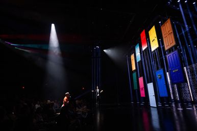 Performer on a dark theater stage under a spotlight engaging the audience, with a playful wall of suspended colorful doors to the right in a large auditorium