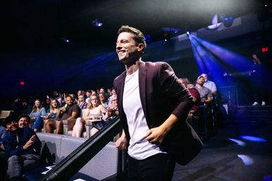 Energetic host with a headset microphone walking up the aisle of a darkened auditorium during a live show, smiling as a seated audience applauds under blue stage lights.