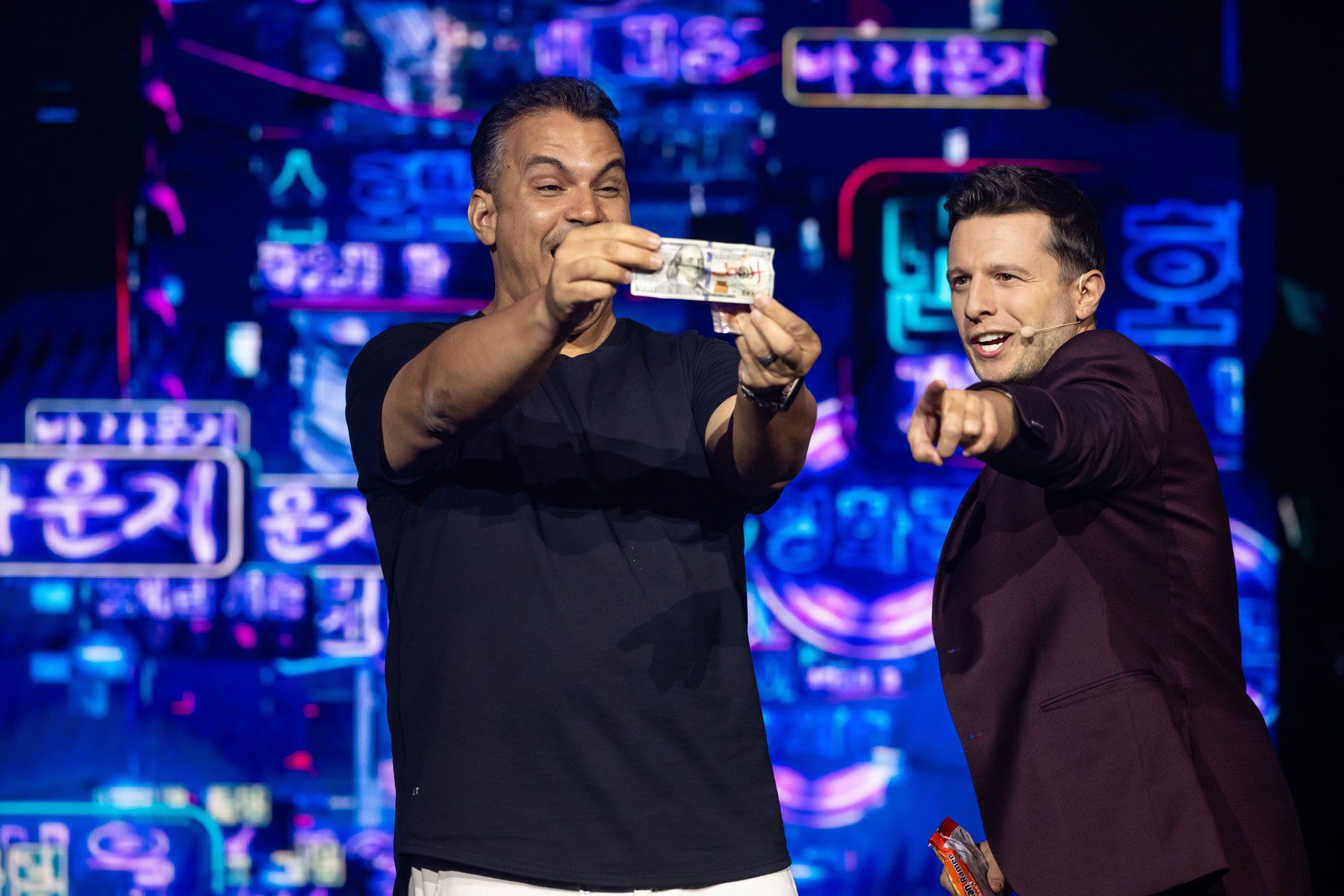 Two men on a neon-lit stage: a smiling participant holds up a banknote while a host in a maroon blazer excitedly points, with vibrant neon Korean signage in the background capturing a lively live-show moment.