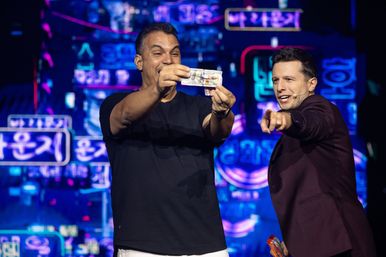 Two men on a neon-lit stage: a smiling participant holds up a banknote while a host in a maroon blazer excitedly points, with vibrant neon Korean signage in the background capturing a lively live-show moment.
