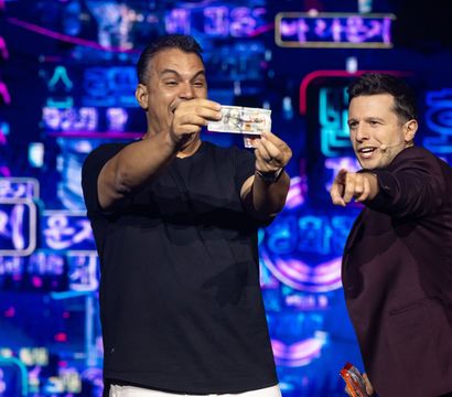 Two men on a neon-lit stage: a smiling participant holds up a banknote while a host in a maroon blazer excitedly points, with vibrant neon Korean signage in the background capturing a lively live-show moment.